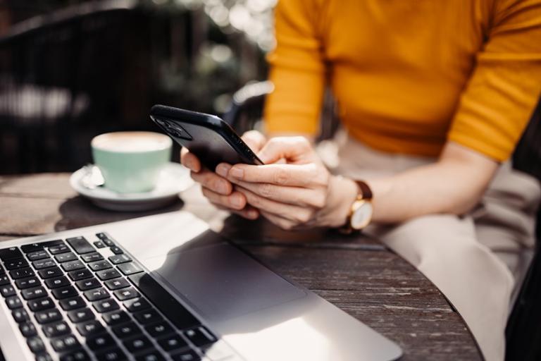 Person's hand holding a smartphone, sat at a table with a laptop